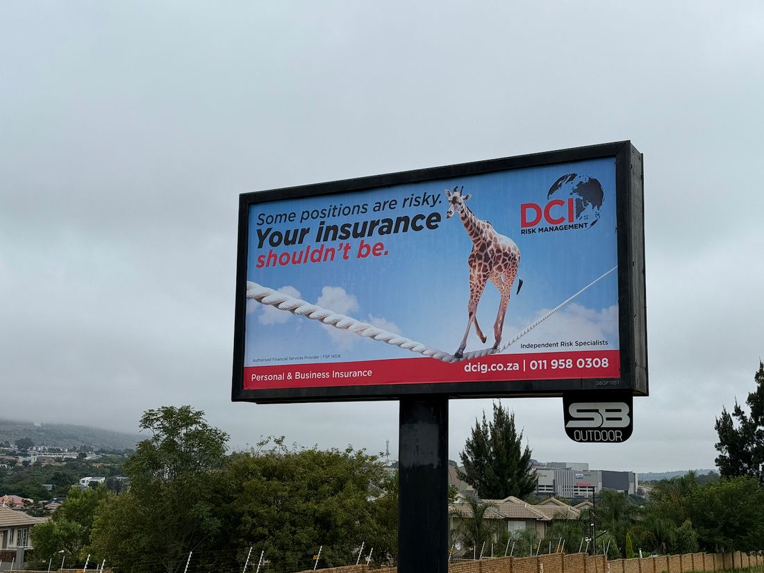 A DCI Risk Management billboard beside a road, a giraffe crossing a tightrope next to the line “Some positions are risky. Your insurance shouldn’t be.”